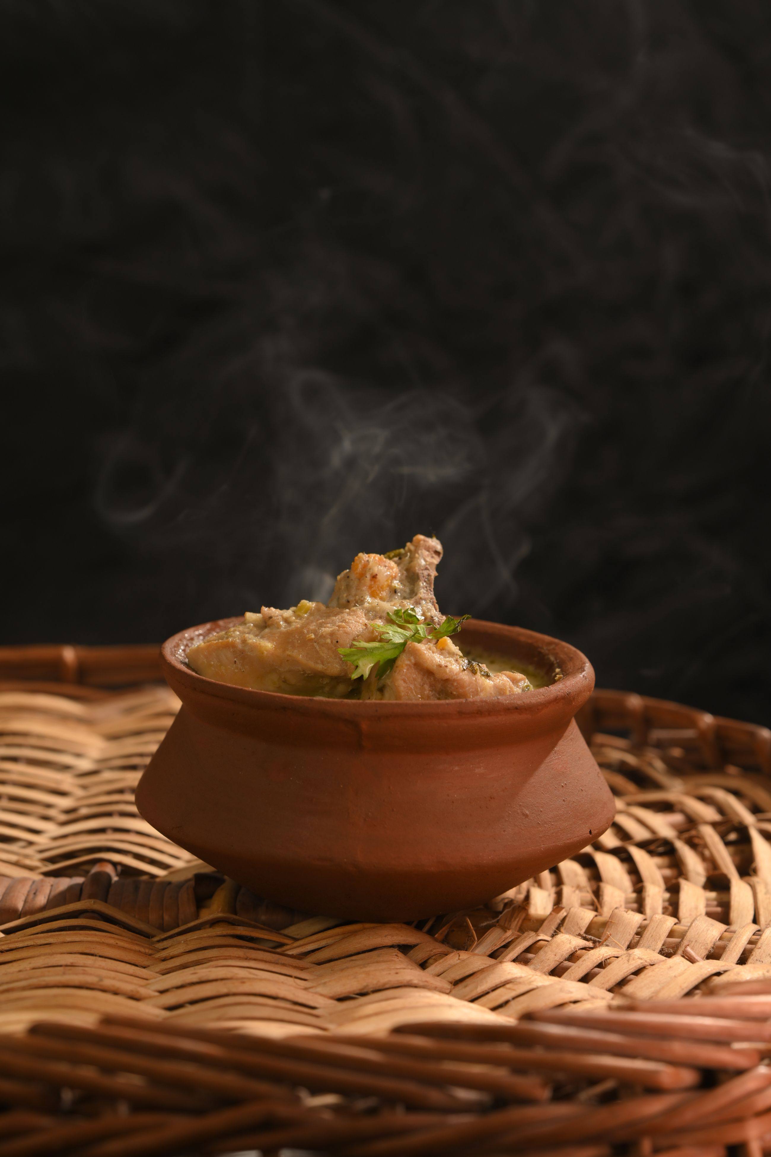 Kerala chicken curry in a small clay pot on a woven cane tray, steam rising against a dark background
