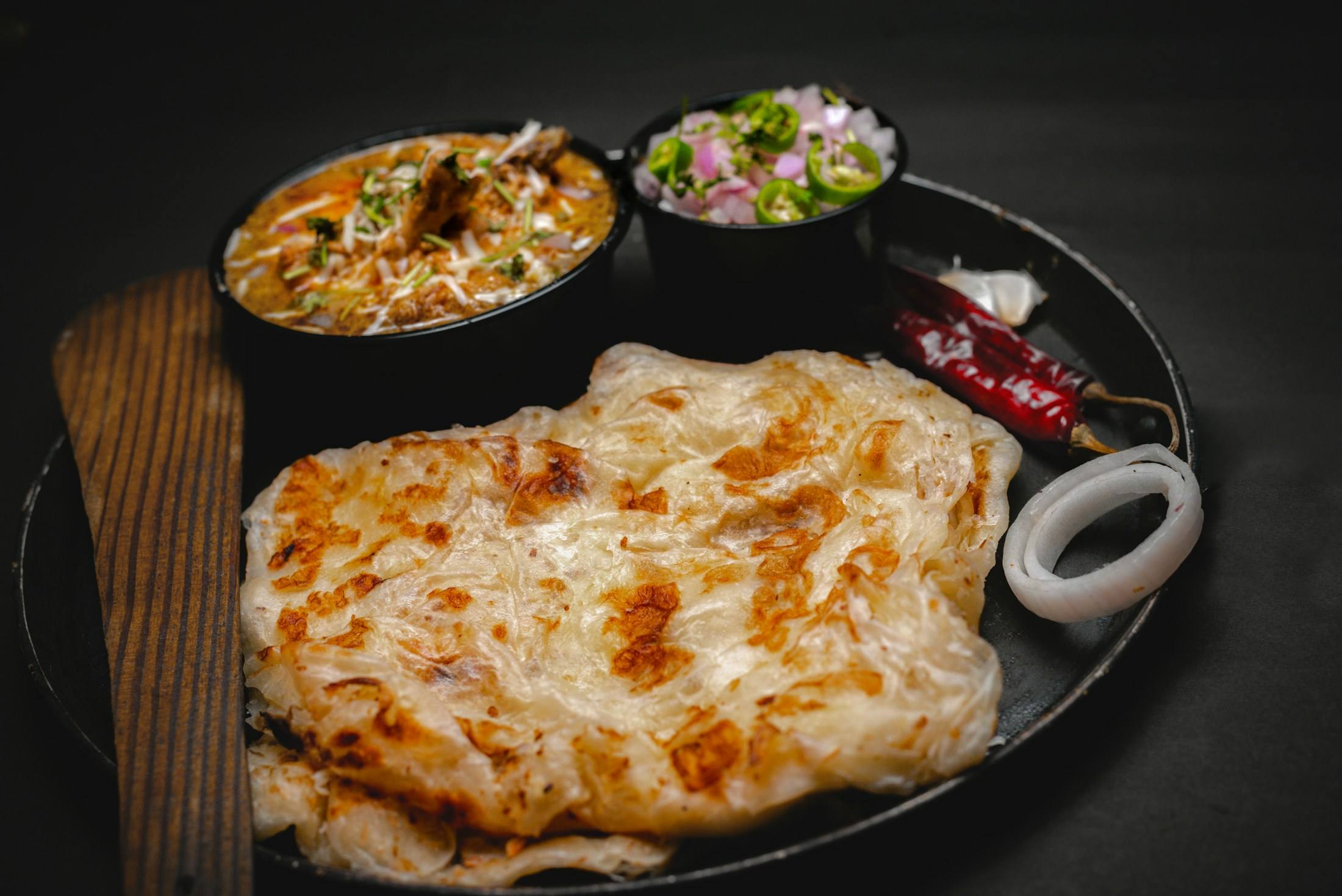 Layered Malabar-style porotta on a black plate with curry, onion and dried chilli