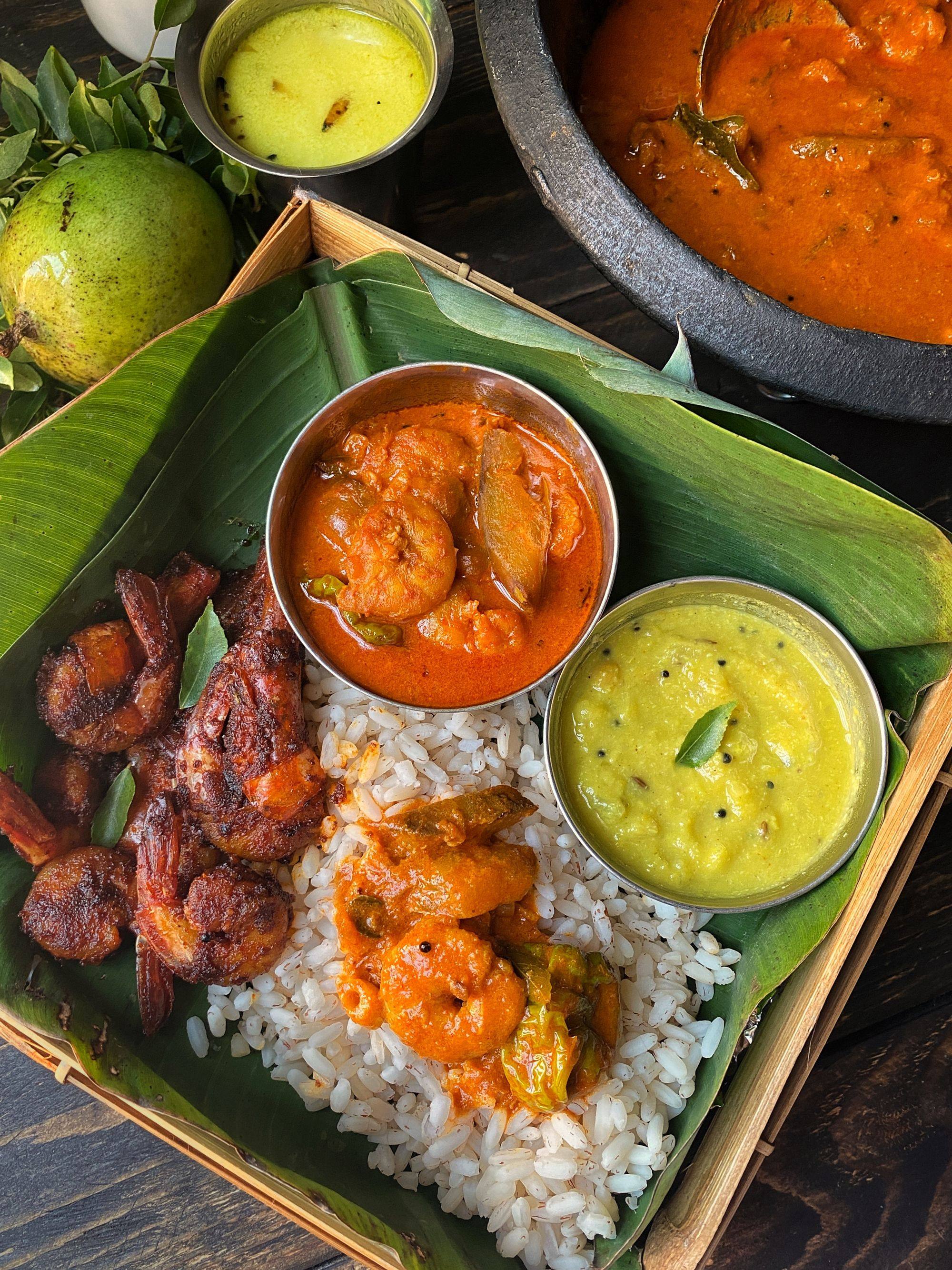 Prawn fry, prawn curry and moru curry over red rice on a banana leaf, a clay pot of fish curry behind